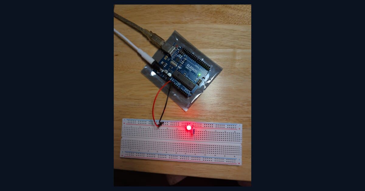 The LED circuit lit on the bench — an ELEGOO UNO R3 (resting on its anti-static bag) feeding a breadboard through a red jumper, with a 220Ω resistor and a glowing red LED.